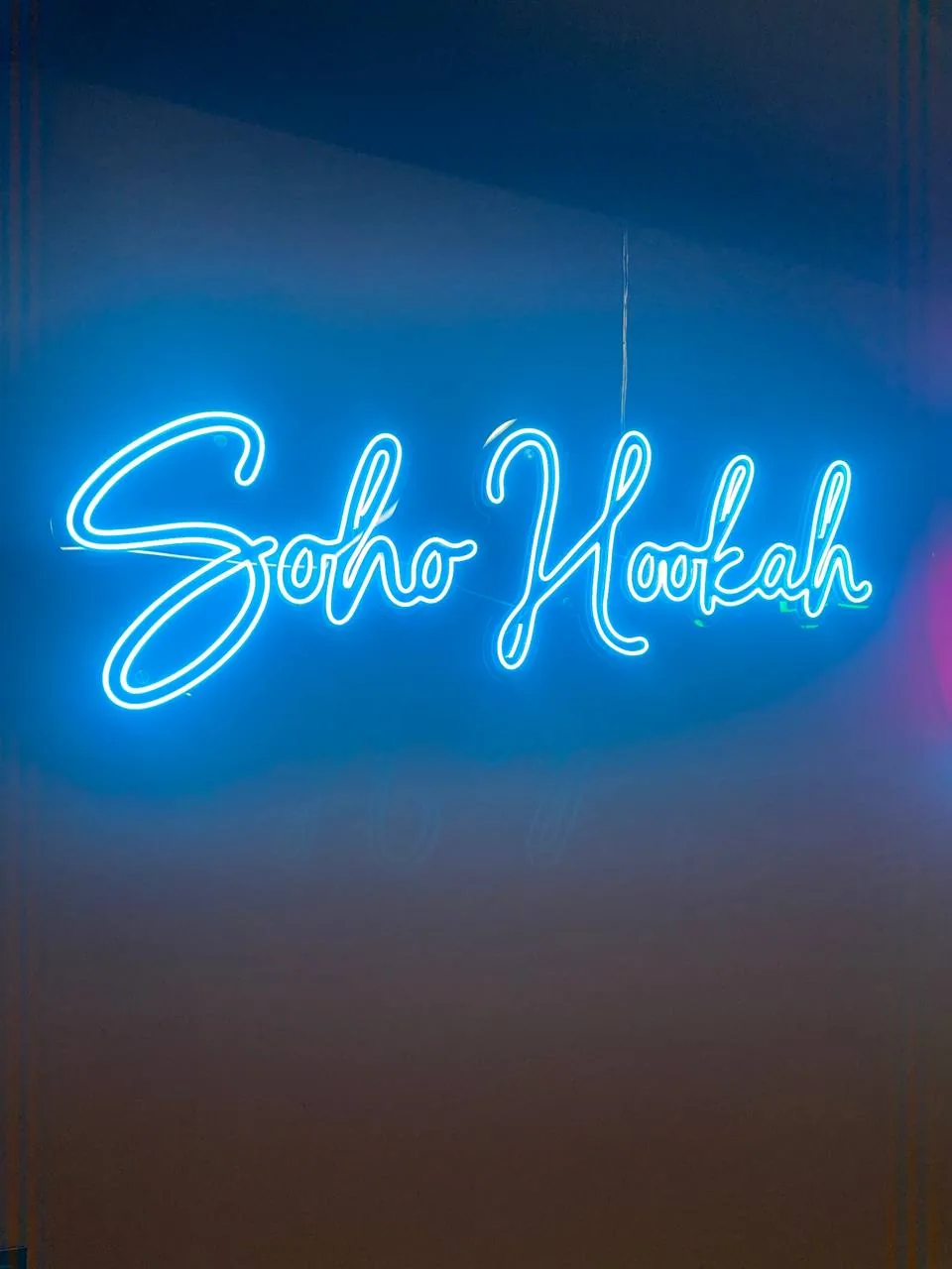 Голубая неоновая надпись Soho Hookah рукописным шрифтом на тёмной стене